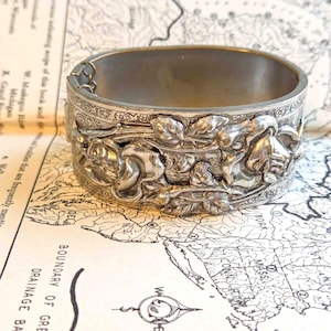 May include: Silver-toned metal cuff bracelet with a floral design. The bracelet features detailed relief carvings of roses and leaves. The clasp is visible on the side. The bracelet is resting on a vintage map.