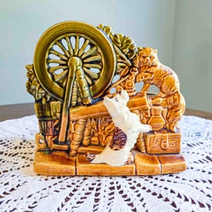 May include: A ceramic figurine featuring a spinning wheel, a dog, and a bear. The wheel is olive green, and the dog is white with brown spots. The bear and base are shades of brown. The figurine has a rustic, vintage aesthetic.