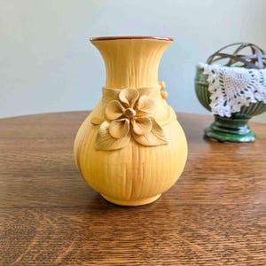 May include: A yellow ceramic vase with a bulbous base and a flared rim. The exterior features a vertical ribbed texture and a prominent three-dimensional sculpted flower with five petals and leaves attached to the front center of the vessel.