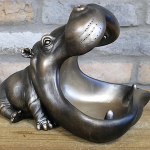 May include: A bronze-coloured hippopotamus figurine with an open mouth, forming a bowl or dish. The hippopotamus is sitting on a wooden surface.