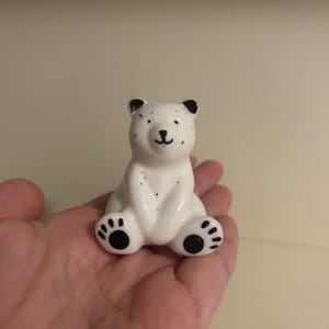 May include: A small white ceramic polar bear figurine with black eyes, nose, and paw prints. The bear is sitting with its paws in front of it.