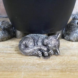Puede incluir: Un conjunto de tres figuras de gato durmiendo de color plateado, utilizadas como soporte de planta para una maceta negra.