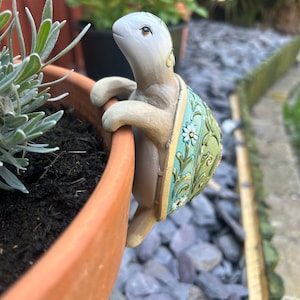 May include: A decorative ceramic turtle figurine with a light grey head and limbs, and a green and cream shell with floral detailing. The turtle is perched on the edge of a terracotta plant pot, adding a decorative touch to a garden.