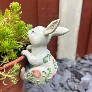 May include: A white ceramic rabbit figurine with floral details climbing a terracotta flower pot. The rabbit has blue eyes and is decorated with green leaves and pink flowers.