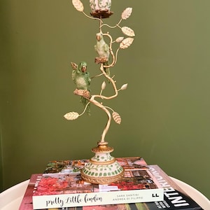 May include: A gold-toned metal candle holder featuring two green parrot figures perched on branches with gold leaves. The base and candle cup are white with green vine patterns. A bright pink taper candle is inserted into the top holder.