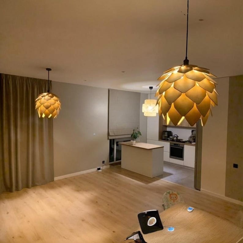 Puede incluir: Tres l&aacute;mparas colgantes modernas iluminan una habitaci&oacute;n espaciosa. La l&aacute;mpara m&aacute;s grande, que se asemeja a una pi&ntilde;a estilizada, cuelga de forma prominente. La habitaci&oacute;n cuenta con suelos de madera clara, paredes neutras y una vista de una cocina.