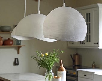 May include: Two white pendant lights with textured, dome-shaped shades hang from the ceiling. The lights are suspended above a kitchen island with a vase of flowers and a bottle of wine.