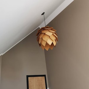 Puede incluir: Una l&aacute;mpara colgante de madera con un dise&ntilde;o &uacute;nico. La l&aacute;mpara est&aacute; suspendida de un cable negro y presenta piezas de madera superpuestas, creando una apariencia en capas y texturizada. La l&aacute;mpara es de color madera natural y c&aacute;lido.