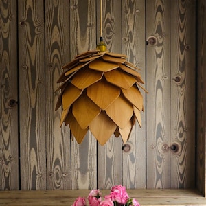 Op de afbeelding: Een houten hanglamp in de vorm van een dennenappel. De lamp is gemaakt van gelaagde, lichtbruine houten bladeren. De lamp hangt aan een goudkleurig snoer tegen een achtergrond van houten planken. Het thema is rustiek en natuurlijk.