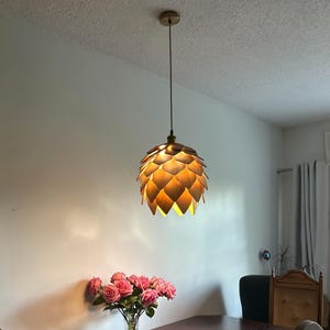 Puede incluir: Una l&aacute;mpara colgante con una cubierta dorada y un cable marr&oacute;n. La l&aacute;mpara tiene una pantalla en capas, en forma de alcachofa, hecha de paneles de madera. La luz cuelga sobre una mesa con un jarr&oacute;n de rosas rosas.