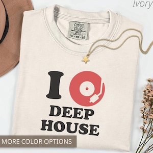 May include: Ivory-colored t-shirt with the text "I Deep House" in black and red. A gold necklace with a star pendant rests on the shirt. The shirt is folded and has a tag that says "Comfort Colors".