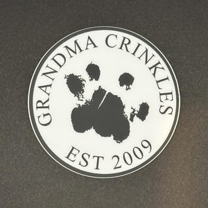 May include: A white circle with black text and a black paw print in the center. The text reads "Grandma Crinckles Est 2009".