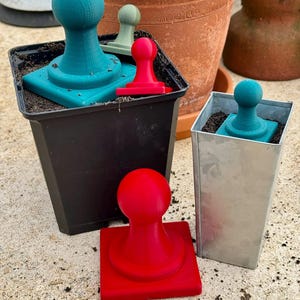 Puede incluir: Una colección de herramientas de jardinería impresas en 3D de colores brillantes. Las herramientas incluyen pisones turquesa, rojos y verdes con bases cuadradas y mangos redondeados. Se muestran en macetas negras y plateadas, sobre una superficie de hormigón.