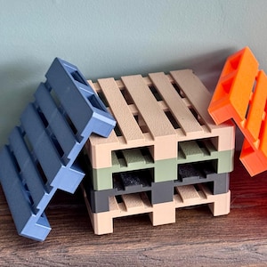 May include: A stack of miniature shipping pallets in various colors including beige, olive green, and dark gray, with one blue pallet leaning against the left side and one bright orange pallet leaning against the right side of the central stack.