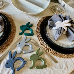 May include: Easter table setting featuring bunny-shaped napkin rings in shades of green, grey, and blue. A white bunny napkin ring decorates a grey napkin. The table is set with black and white plates, woven placemats, and cutlery.