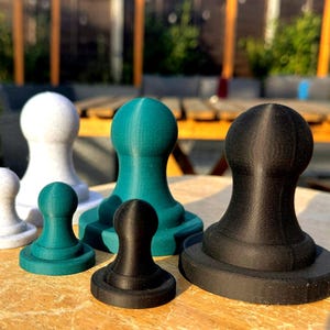 May include: A collection of six 3D-printed objects in various sizes and colors, including white, teal, and black. Each object has a rounded top and a circular base, resembling chess pieces or decorative items. They are arranged on a wooden surface.