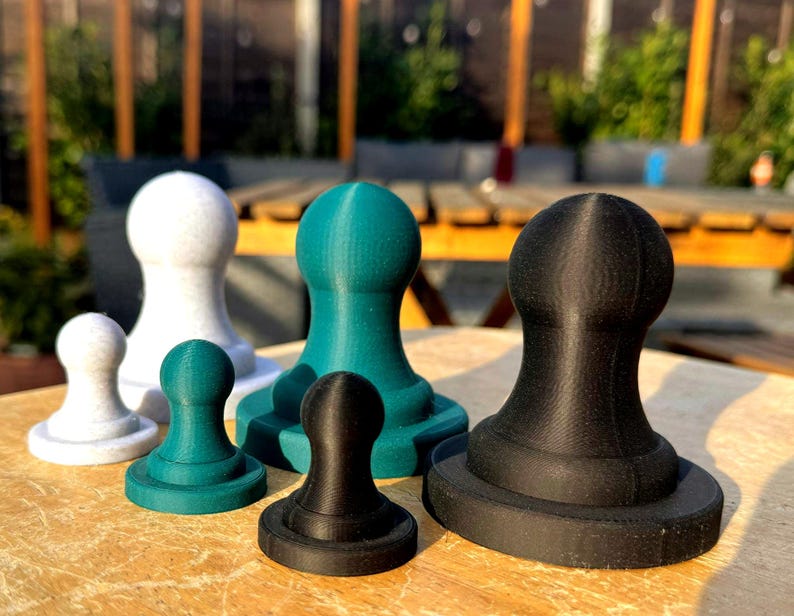 May include: A collection of six 3D-printed objects in various sizes and colors, including white, teal, and black. Each object has a rounded top and a circular base, resembling chess pieces or decorative items. They are arranged on a wooden surface.