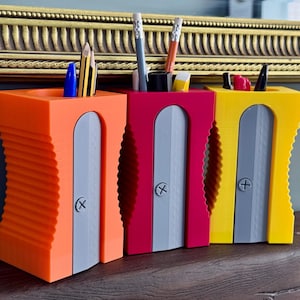 May include: Three pencil holders shaped like large pencil sharpeners in orange, magenta, and yellow. Each holder features a gray front panel with a screw detail and a ridged side texture. Various pens and pencils are stored inside the top openings.