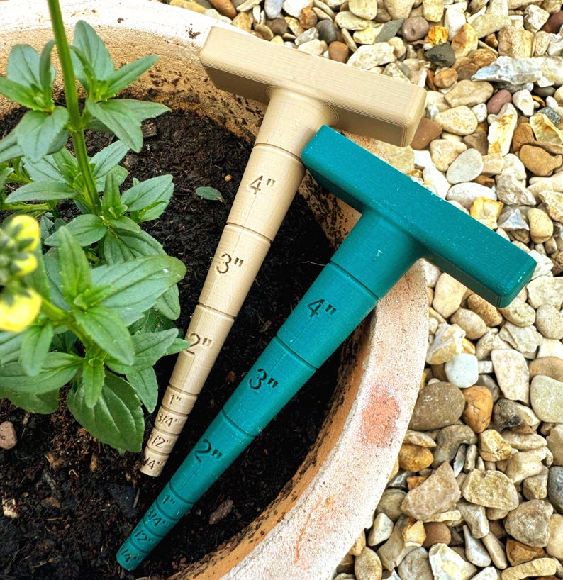 May include: Two T-shaped plant water indicators. One is beige, the other is teal. Both have measurement markings in inches, from 1/2" to 4". They are placed in a terracotta pot with soil and plants, surrounded by small rocks.