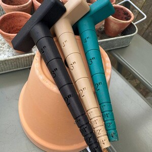 May include: Three T-shaped gardening tools in black, tan, and teal, each marked with inch measurements from 1/4" to 4". The tools are resting on a terracotta pot, with other pots and a metal tray in the background.