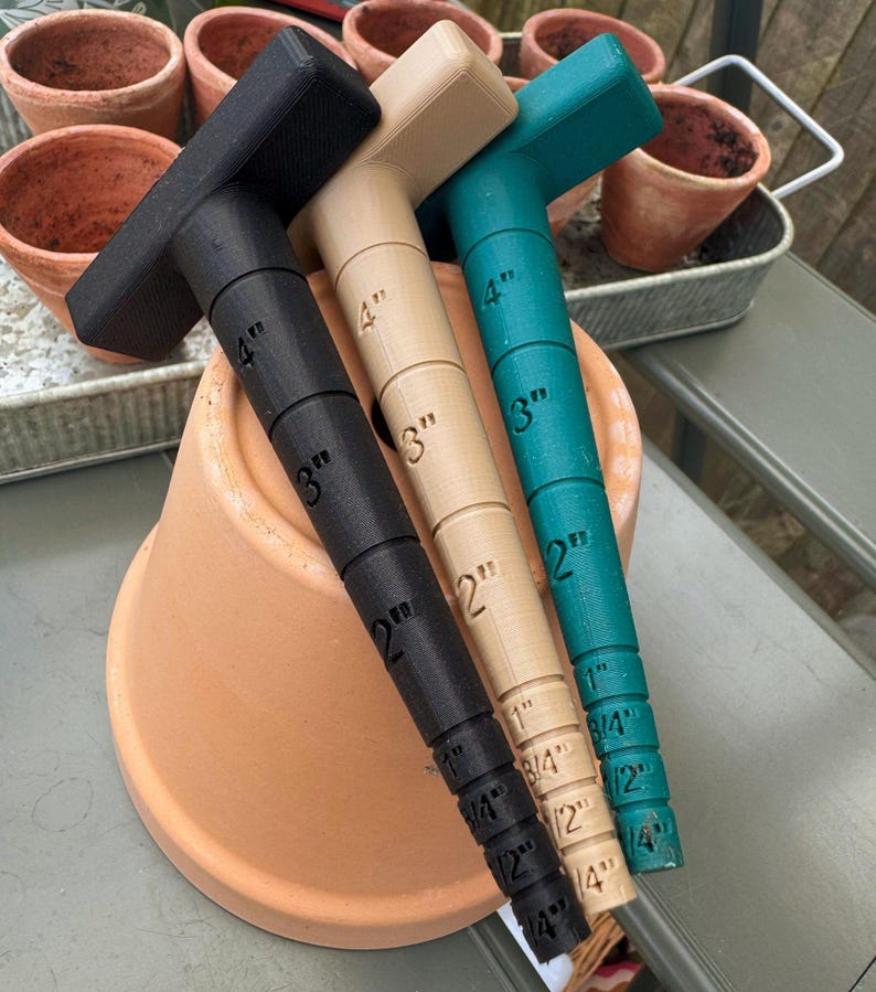 May include: Three T-shaped gardening tools in black, tan, and teal, each marked with inch measurements from 1/4" to 4". The tools are resting on a terracotta pot, with other pots and a metal tray in the background.