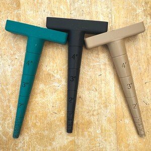 May include: Three T-shaped tools in teal, black, and tan, each with a graduated conical body. The tools are marked with inch measurements from 1/4" to 4". The tools are likely used for measuring or marking.