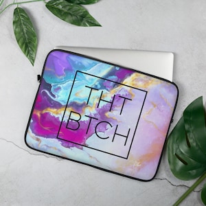 May include: A laptop sleeve with a colorful abstract design featuring shades of purple, pink, blue, and gold. The text "THAT BTCH" is printed in a black square on the sleeve.