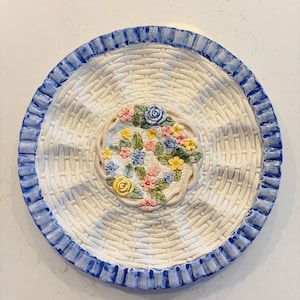 Peut inclure: Une assiette de service ronde en céramique blanche avec un motif de panier tressé. L'assiette présente un bord bleu et un arrangement floral central aux couleurs pastel. Idéale pour servir des hors-d'œuvre ou des desserts.