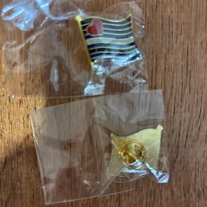 Puede incluir: Dos pines de esmalte con forma de bandera. Un pin presenta un diseño con rayas azules y doradas, un borde negro y un corazón rojo. El otro pin es de color dorado con un pasador.