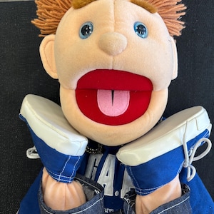 Può includere: Un pupazzo di peluche con carnagione chiara, occhi blu e capelli arancioni. Il pupazzo ha la bocca aperta con l'interno rosso e la lingua rosa. Indossa una camicia blu e jeans, con scarpe da ginnastica blu e bianche.