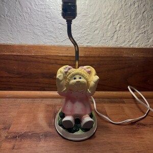Op de afbeelding: Een vintage Cabbage Patch Kids lamp met een keramieken beeldje van een meisje in een roze jurk. De lamp heeft een messingkleurige hals en een witte voet met groene accenten. De lamp wordt verlicht door een heldere gloeilamp.