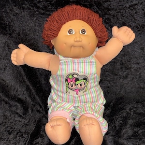 May include: A vintage doll with a soft, fabric body and a vinyl face. The doll has red yarn hair, large eyes, and a smiling expression. It wears a striped jumpsuit with a graphic design and pink accents. The doll's arms are outstretched.