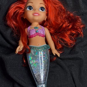 May include: A red-haired mermaid doll with a purple and pink seashell top and a glittery turquoise tail.