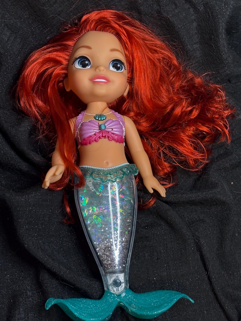 May include: A red-haired mermaid doll with a purple and pink seashell top and a glittery turquoise tail.
