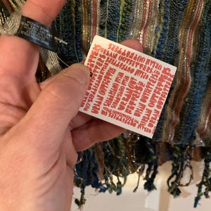 K&ouml;nnte beinhalten: Ein wei&szlig;es Etikett mit rotem Text, auf dem mehrmals "URBAN OUTFITTERS" steht. Das Etikett ist an einem mehrfarbigen gestreiften Schal mit Fransen befestigt.