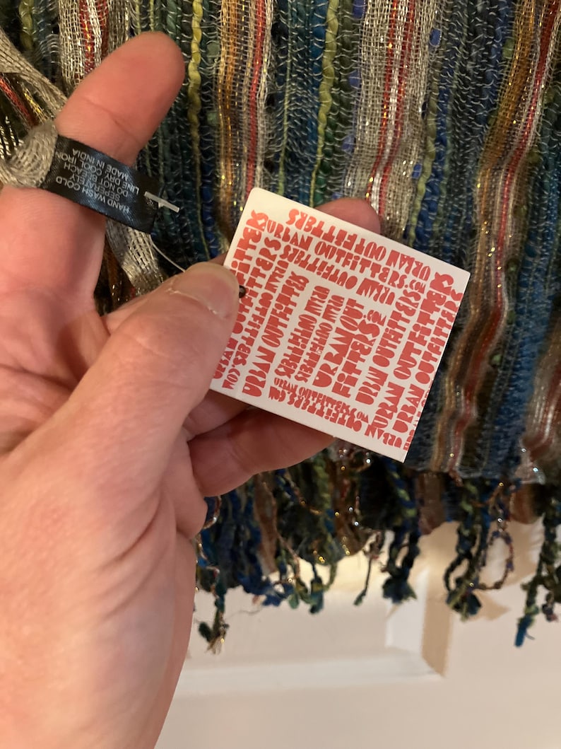 K&ouml;nnte beinhalten: Ein wei&szlig;es Etikett mit rotem Text, auf dem mehrmals "URBAN OUTFITTERS" steht. Das Etikett ist an einem mehrfarbigen gestreiften Schal mit Fransen befestigt.
