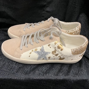 May include: A pair of low-top sneakers with a white base and brown cowhide print. The sneakers have a gold star with silver glitter on the side and a gold glitter heel.