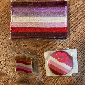Peut inclure: Trois articles de fierté lesbienne : un patch en tissu à rayures rouges, blanches et violettes, une épingle dorée avec un drapeau à rayures rouges, blanches et violettes et un bouton rond avec un fond à rayures rouges, blanches et violettes et une empreinte de lèvres rouges.