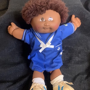 Może przedstawiać: Lalka Cabbage Patch Kid z brązowymi kręconymi włosami, brązowymi oczami i różowymi policzkami. Lalka ma na sobie niebieski strój marynarski z białym kołnierzykiem i białą kokardką. Lalka ma również białe skarpetki i beżowe buty sportowe z niebieskimi paskami.