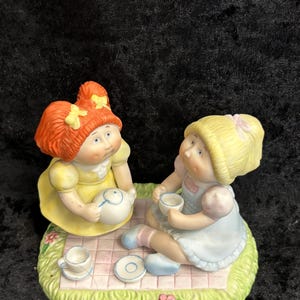 Op de afbeelding: Een keramieken beeldje met twee poppen die een theeparty houden. De ene pop draagt een gele jurk, de andere een blauwe jurk. Ze zitten op een roze en witte geblokte deken op een groene basis met bloemen.