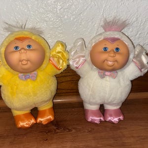 May include: Two plush dolls, one yellow and one white, in animal costumes. The yellow doll is dressed as a chick with orange feet and a bow tie. The white doll is dressed as a lamb with pink feet and a bow tie. Both dolls have blue eyes and a smiling expression.