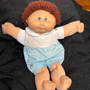 Könnte beinhalten: Eine Vintage-Puppe mit einem weichen Stoffkörper. Die Puppe hat einen hellen Teint, braunes Garnhaar und trägt ein weißes Hemd mit Kragen und hellblaue Shorts. Die Arme und Beine der Puppe sind ausgestreckt.