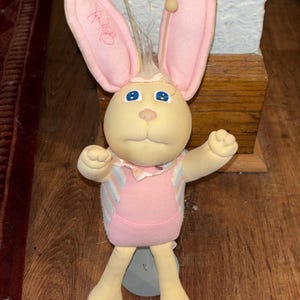 Puede incluir: Un muñeco de peluche con forma de conejo de tela color crema, grandes orejas forradas de rosa y ojos azules. Presenta dos antenas con puntas redondas en la cabeza, un lazo rosa en el cuello y un conjunto a rayas rosas y azul claro. Firma en la oreja.
