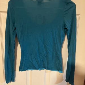 May include: A teal blue long-sleeved shirt with a scoop neck and a slightly ruffled hem.