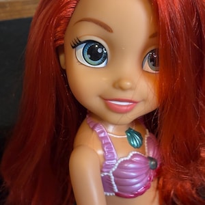 May include: A red-haired doll with blue and green eyes, wearing a pink seashell top and a green seashell bottom. The doll is smiling and has a seashell necklace.