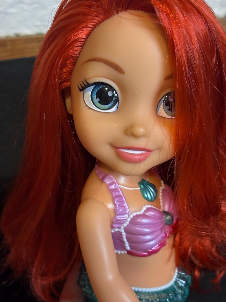 May include: A red-haired doll with blue and green eyes, wearing a pink seashell top and a green seashell bottom. The doll is smiling and has a seashell necklace.