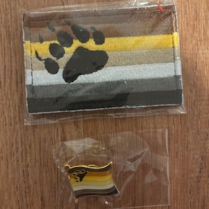 Peut inclure: Un écusson et une épingle du drapeau de la fierté des ours. L'écusson est rectangulaire avec des rayures horizontales marron, jaune, grise et noire, et une empreinte de patte noire. L'épingle a le même design en plus petit.