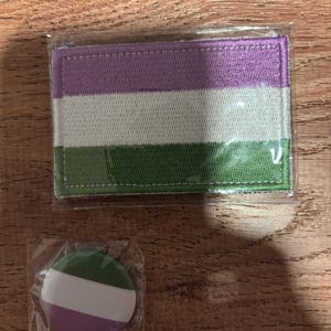Peut inclure: Un écusson rectangulaire et un bouton rond, tous deux arborant le drapeau de la fierté genderqueer. L'écusson présente des rayures horizontales violettes, blanches et vertes. Le bouton reprend ce motif en cercle. Les deux articles sont dans un emballage transparent.