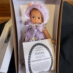 Puede incluir: Una muñeca de porcelana llamada Patsy, que lleva un vestido lavanda y un gorro de encaje blanco con una flor rosa. La muñeca está en una caja blanca con un certificado de autenticidad que dice "Certificado de Autenticidad Patsy Spring Fling Patsy hecho exclusivamente para Ashton-Drake Galleries".