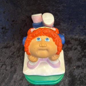 May include: A vintage Cabbage Patch Kids figurine. The doll has orange hair, blue eyes, and a tan complexion. It is wearing a pink and white outfit and is resting on a green and white base. The figurine is a collectible item.